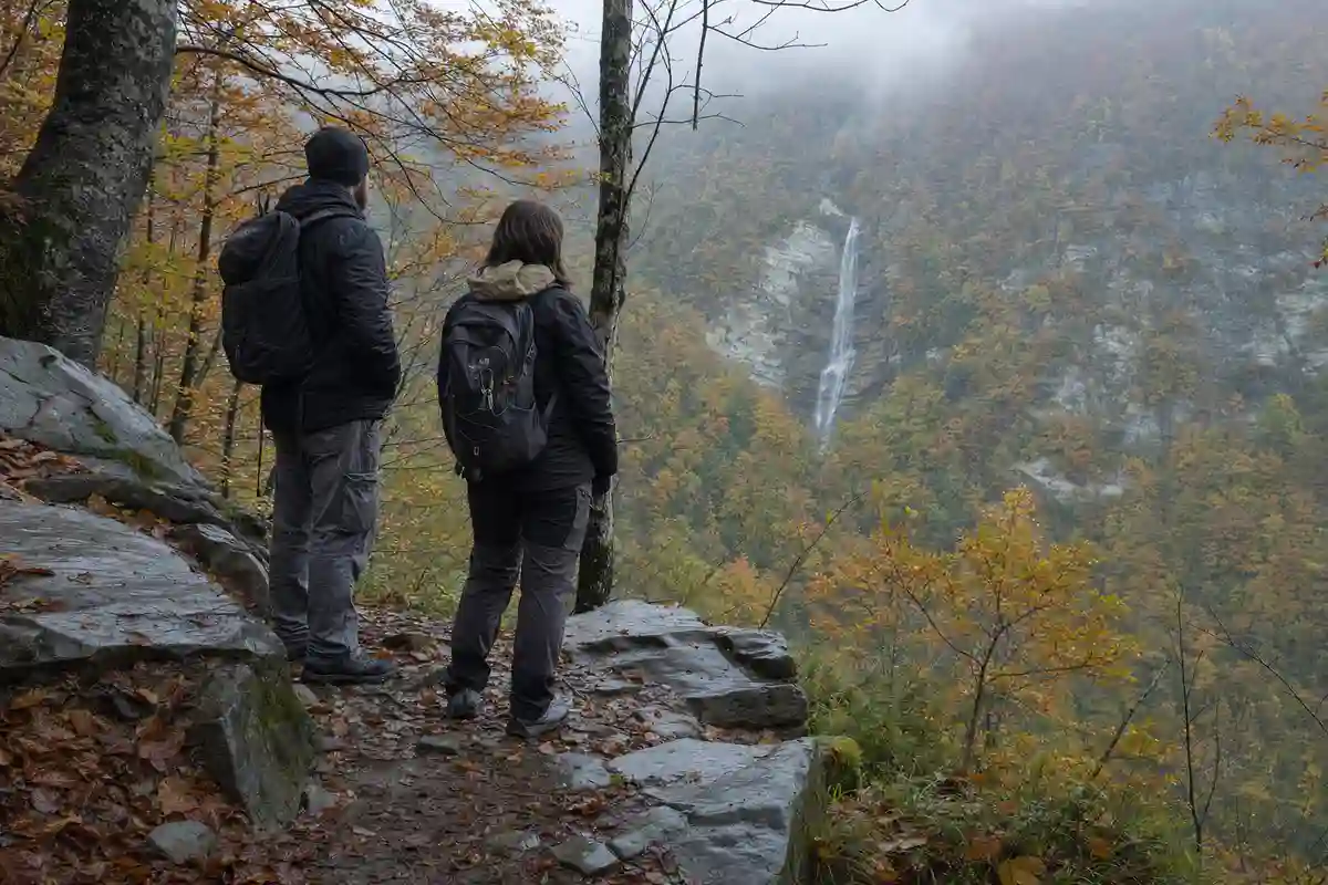 Due escursionisti su un sentiero roccioso osservano una cascata lontana tra boschi autunnali e nebbia