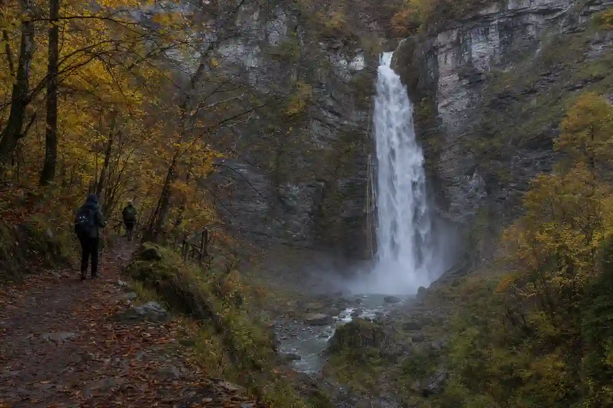 Cascata alta tra pareti rocciose in un bosco autunnale, vista dal sentiero con due escursionisti in lontananza