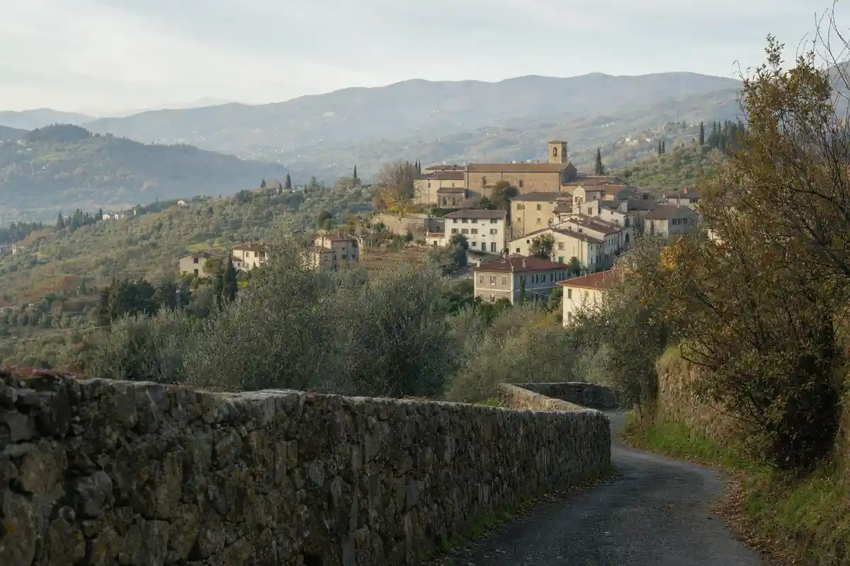 Strada di campagna con muretto in pietra che conduce a un borgo collinare tra olivi e colline toscane