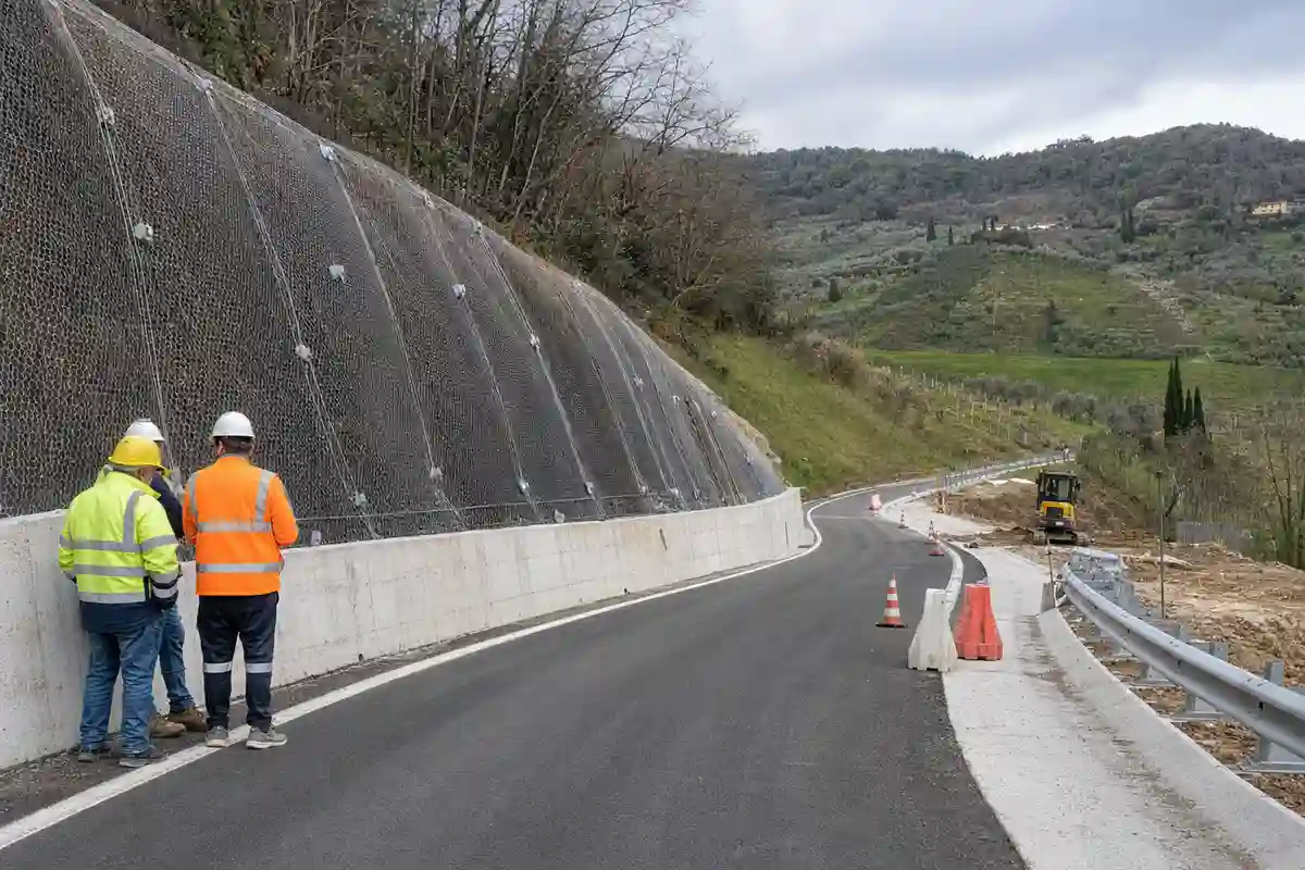 Cantiere su strada collinare con rete sul versante, new jersey e coni; tecnici in giubbotto alta visibilità