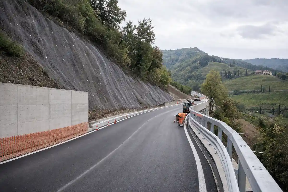 Strada provinciale in collina con asfalto nuovo, rete sul versante, coni e operaio al lavoro su guardrail