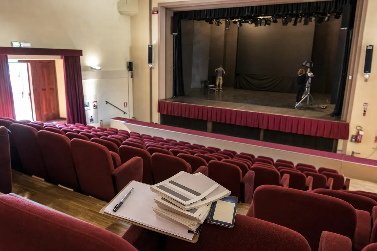 Platea di un piccolo teatro con poltrone rosse e palco con sipario scuro, fogli e clipboard in primo piano